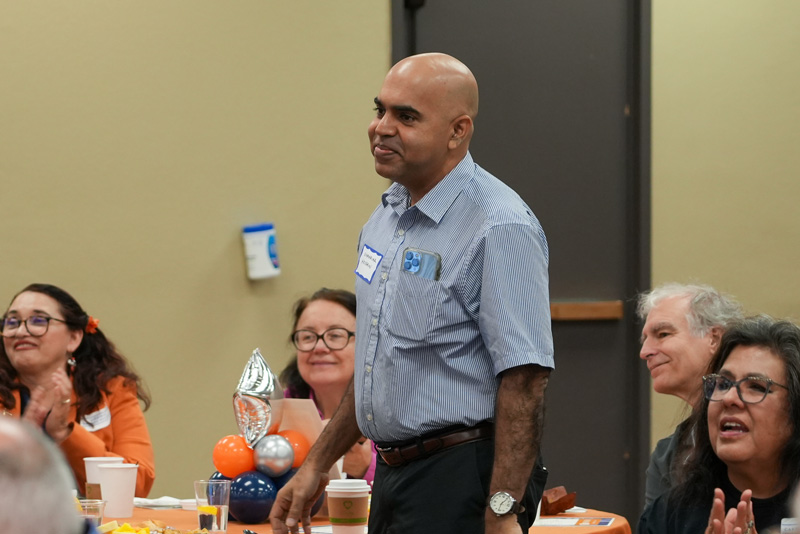 Individual speaks from a podium during a UTEP Engineering homecoming gathering, with a large orange College of Engineering balloon decoration and a projected portrait displayed on the presentation screen behind the speaker.